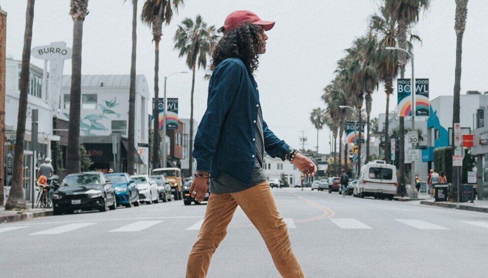 A woman walking across a crosswalk in a sunny urban area with palm trees and shops.