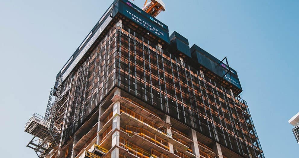 Modern skyscraper under construction with scaffolding and cranes against blue sky.