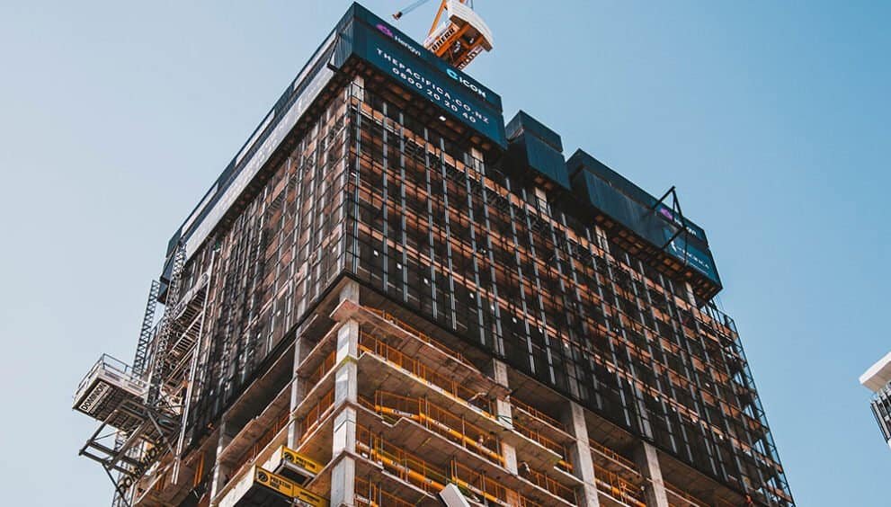 Modern skyscraper under construction with scaffolding and cranes against blue sky.