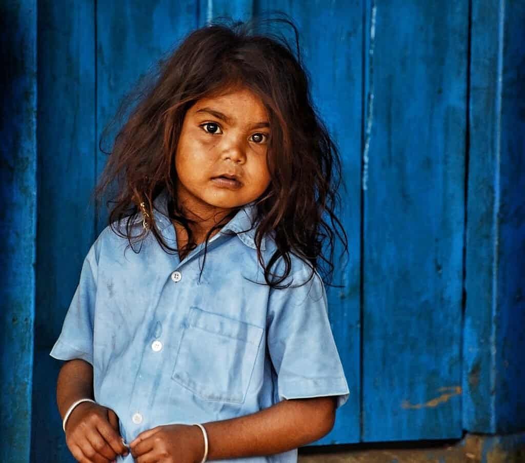 Hopeful young girl in traditional clothing stands in front of a blue wooden background, symbolizing resilience and community.