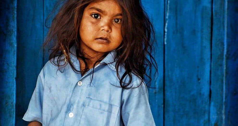 Hopeful young girl in traditional clothing stands in front of a blue wooden background, symbolizing resilience and community.