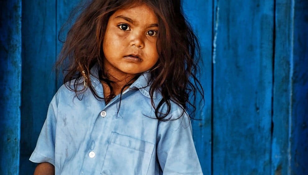 Hopeful young girl in traditional clothing stands in front of a blue wooden background, symbolizing resilience and community.