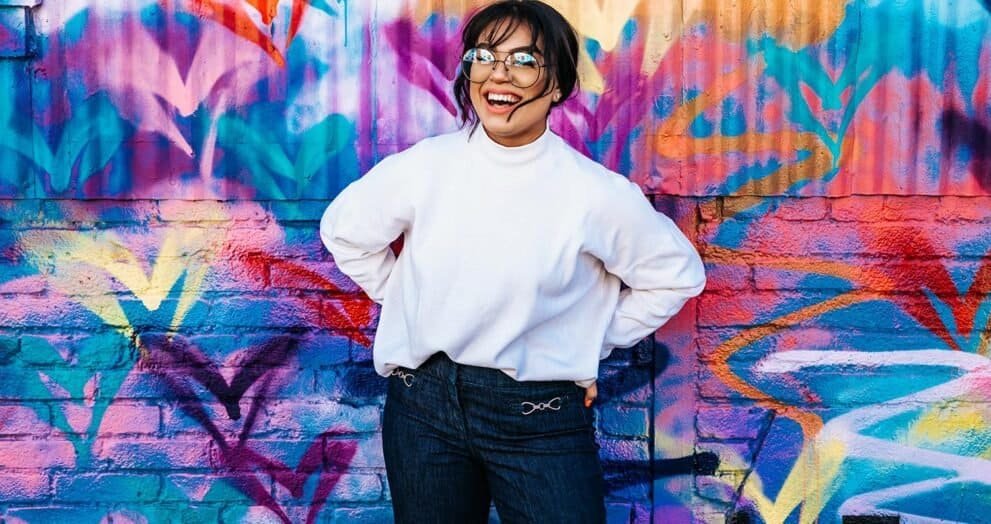 Colorful graffiti mural featuring a smiling woman in casual white sweatshirt and dark jeans.